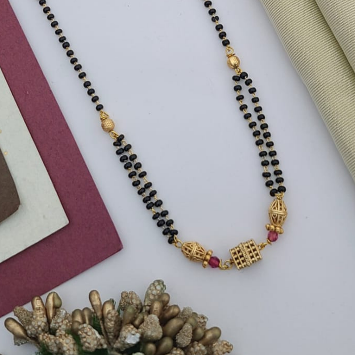 Gold-plated mangalsutra with dual black bead chains and intricate golden pendant design, displayed on a flat lay with fabric and decorative props – traditional jewellery by RC Imitation Jewellery.