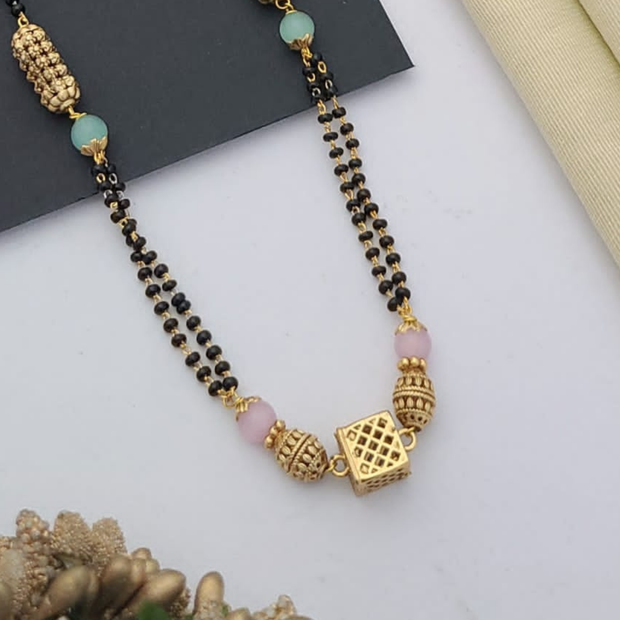 Gold-plated mangalsutra with dual black bead chains and intricate golden pendant design, displayed on a flat lay with fabric and decorative props – traditional jewellery by RC Imitation Jewellery.