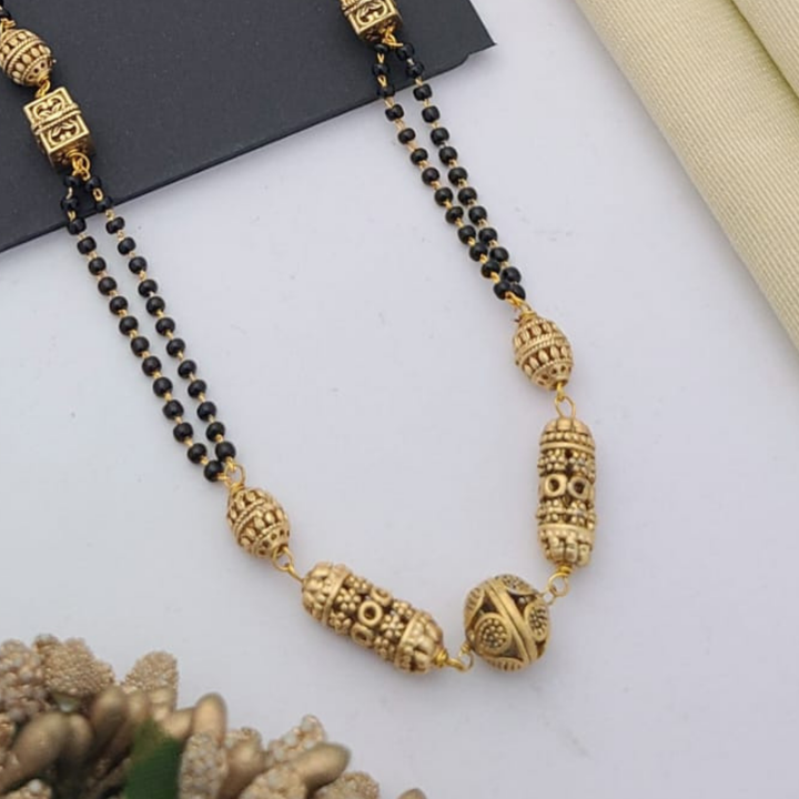 Gold-plated mangalsutra with dual black bead chains and intricate golden pendant design, displayed on a flat lay with fabric and decorative props – traditional jewellery by RC Imitation Jewellery.