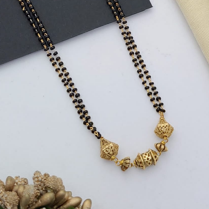 Gold-plated mangalsutra with dual black bead chains and intricate golden pendant design, displayed on a flat lay with fabric and decorative props – traditional jewellery by RC Imitation Jewellery.