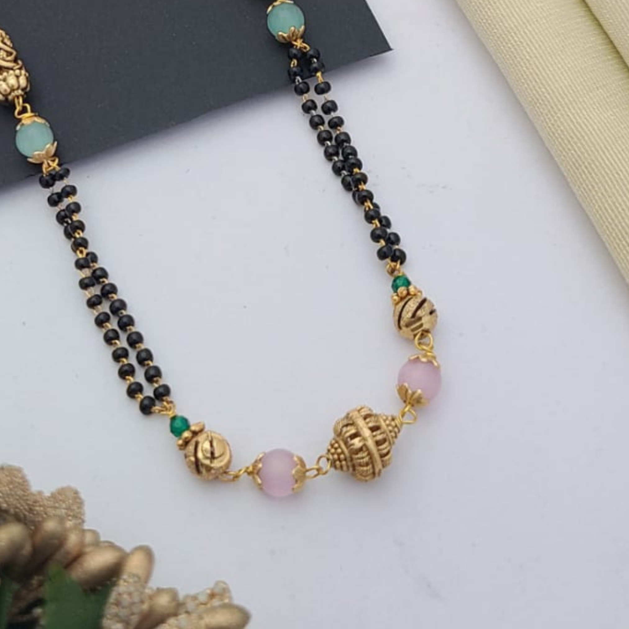 Gold-plated mangalsutra with dual black bead chains and intricate golden pendant design, displayed on a flat lay with fabric and decorative props – traditional jewellery by RC Imitation Jewellery.