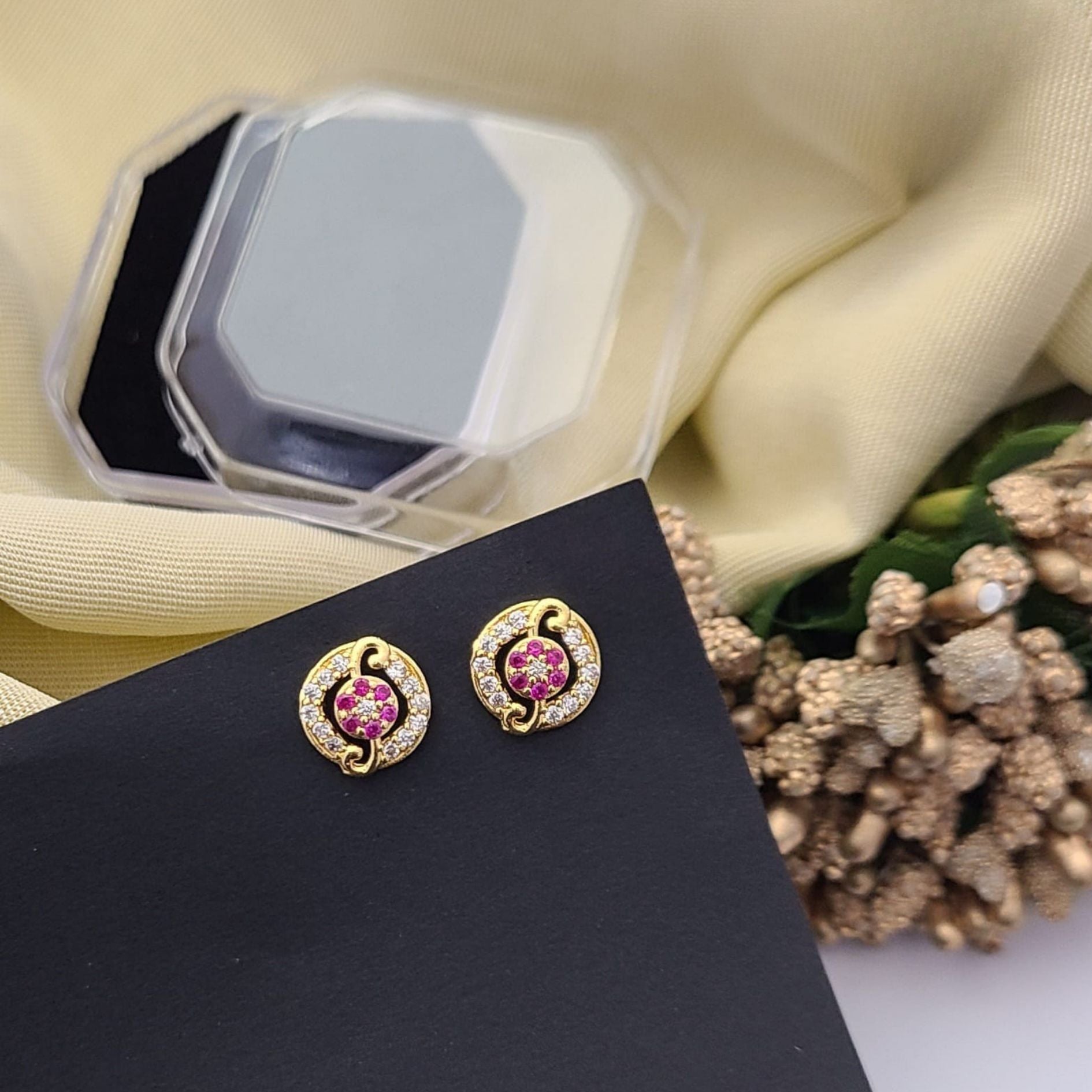 Gold plated round stud earrings with pink floral center and sparkling white stones, displayed on a black card with a decorative background