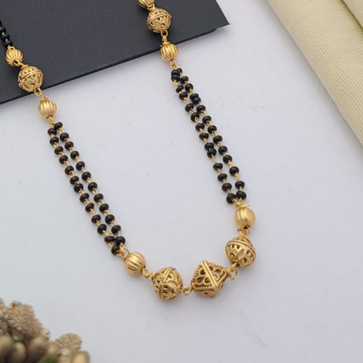 Gold-plated mangalsutra with dual black bead chains and intricate golden pendant design, displayed on a flat lay with fabric and decorative props – traditional jewellery by RC Imitation Jewellery.