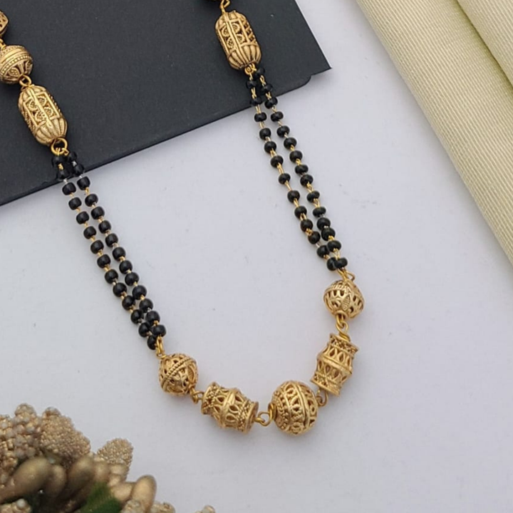 Gold-plated mangalsutra with dual black bead chains and intricate golden pendant design, displayed on a flat lay with fabric and decorative props – traditional jewellery by RC Imitation Jewellery.