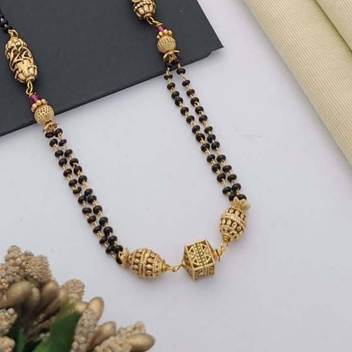 Gold-plated mangalsutra with dual black bead chains and intricate golden pendant design, displayed on a flat lay with fabric and decorative props – traditional jewellery by RC Imitation Jewellery.