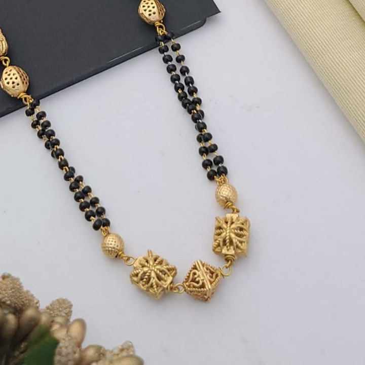 Gold-plated mangalsutra with dual black bead chains and intricate golden pendant design, displayed on a flat lay with fabric and decorative props – traditional jewellery by RC Imitation Jewellery.