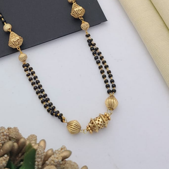 Gold-plated mangalsutra with dual black bead chains and intricate golden pendant design, displayed on a flat lay with fabric and decorative props – traditional jewellery by RC Imitation Jewellery.
