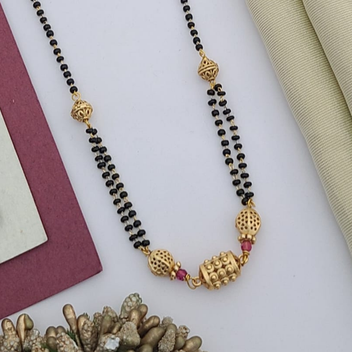 Gold-plated mangalsutra with dual black bead chains and intricate golden pendant design, displayed on a flat lay with fabric and decorative props – traditional jewellery by RC Imitation Jewellery.