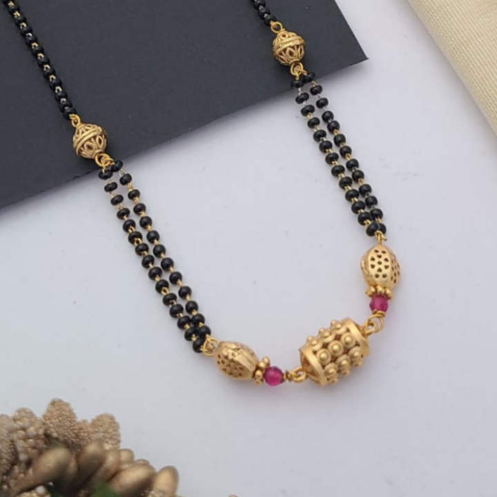 Gold-plated mangalsutra with dual black bead chains and intricate golden pendant design, displayed on a flat lay with fabric and decorative props – traditional jewellery by RC Imitation Jewellery.