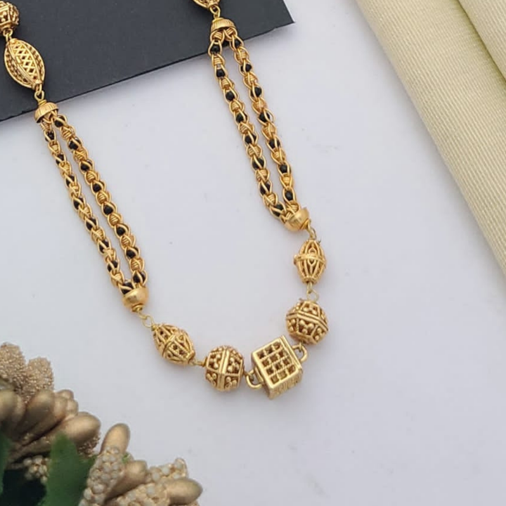 Gold-plated mangalsutra with dual black bead chains and intricate golden pendant design, displayed on a flat lay with fabric and decorative props – traditional jewellery by RC Imitation Jewellery.
