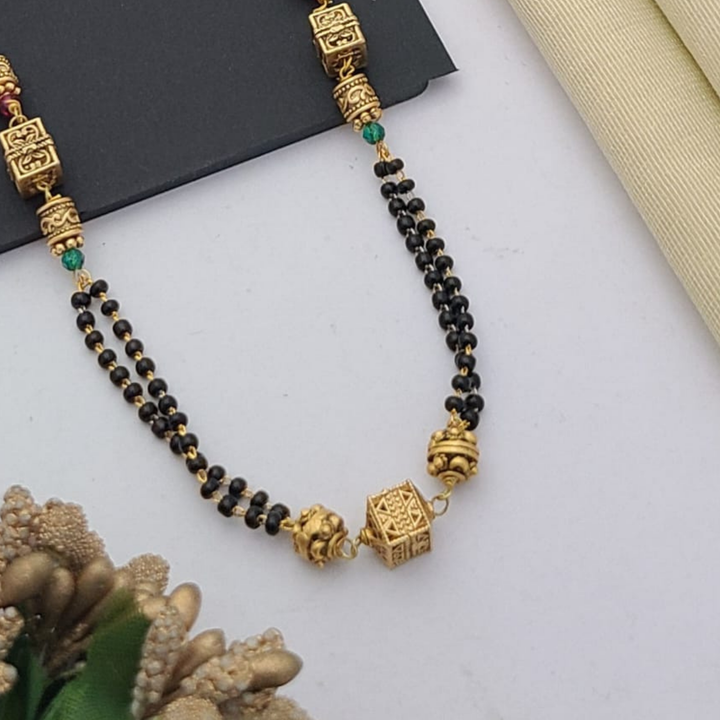 Gold-plated mangalsutra with dual black bead chains and intricate golden pendant design, displayed on a flat lay with fabric and decorative props – traditional jewellery by RC Imitation Jewellery.