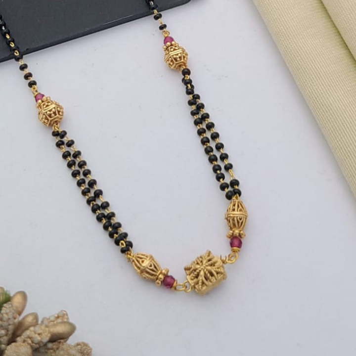 Gold-plated mangalsutra with dual black bead chains and intricate golden pendant design, displayed on a flat lay with fabric and decorative props – traditional jewellery by RC Imitation Jewellery.