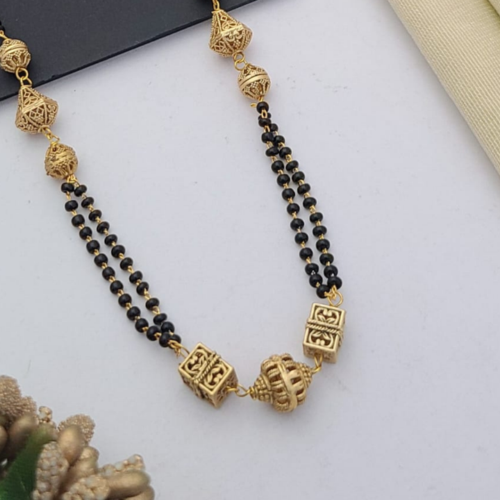 Gold-plated mangalsutra with dual black bead chains and intricate golden pendant design, displayed on a flat lay with fabric and decorative props – traditional jewellery by RC Imitation Jewellery.