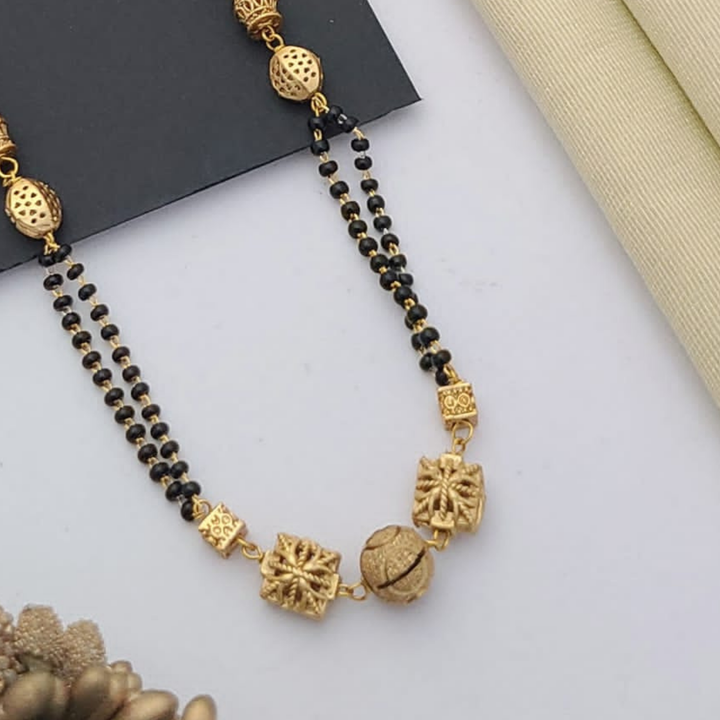 Gold-plated mangalsutra with dual black bead chains and intricate golden pendant design, displayed on a flat lay with fabric and decorative props – traditional jewellery by RC Imitation Jewellery.