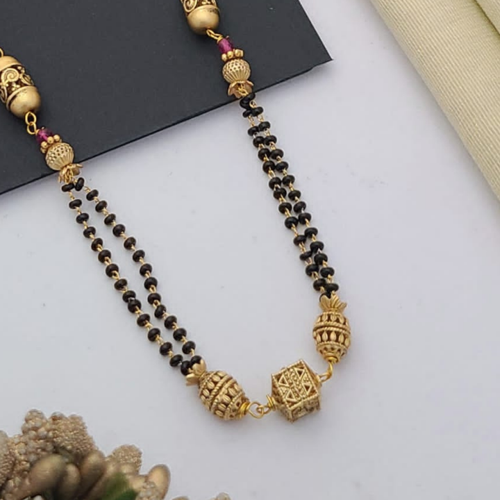 Gold-plated mangalsutra with dual black bead chains and intricate golden pendant design, displayed on a flat lay with fabric and decorative props – traditional jewellery by RC Imitation Jewellery.