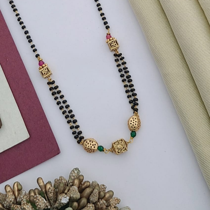 Gold-plated mangalsutra with dual black bead chains and intricate golden pendant design, displayed on a flat lay with fabric and decorative props – traditional jewellery by RC Imitation Jewellery.