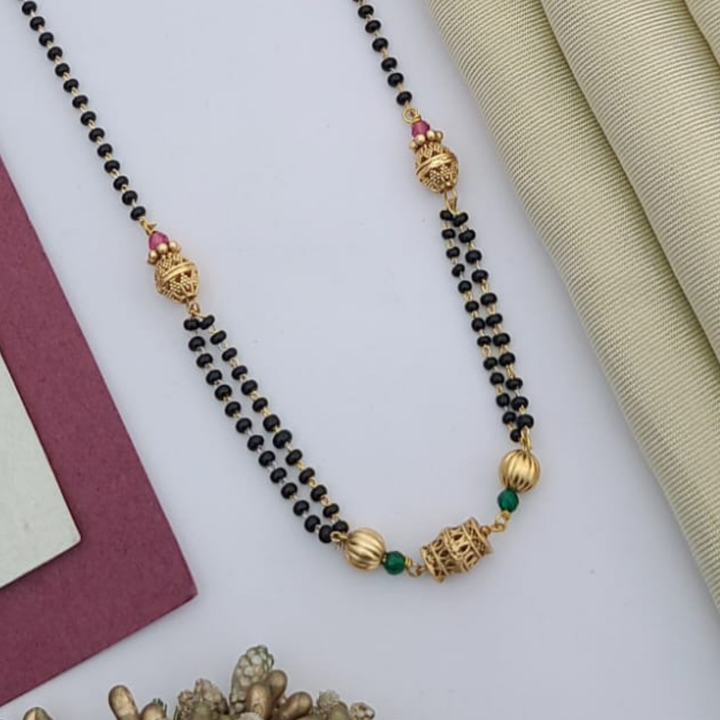 Gold-plated mangalsutra with dual black bead chains and intricate golden pendant design, displayed on a flat lay with fabric and decorative props – traditional jewellery by RC Imitation Jewellery.