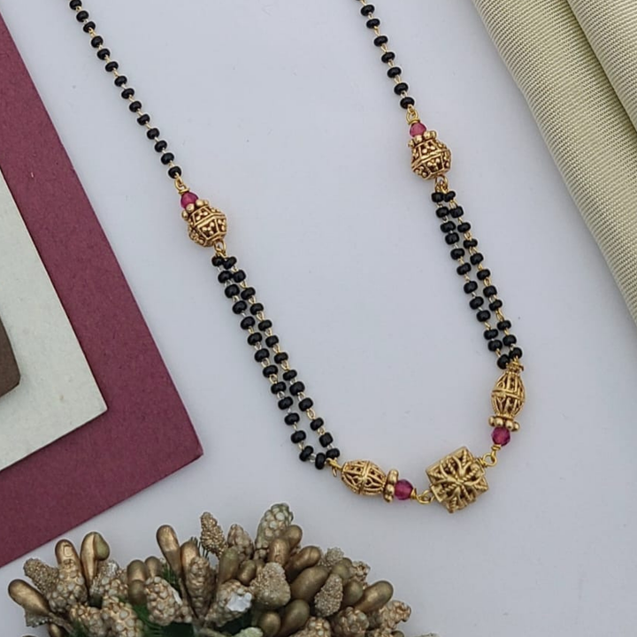 Gold-plated mangalsutra with dual black bead chains and intricate golden pendant design, displayed on a flat lay with fabric and decorative props – traditional jewellery by RC Imitation Jewellery.