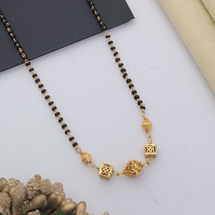 Gold-plated mangalsutra with dual black bead chains and intricate golden pendant design, displayed on a flat lay with fabric and decorative props – traditional jewellery by RC Imitation Jewellery.